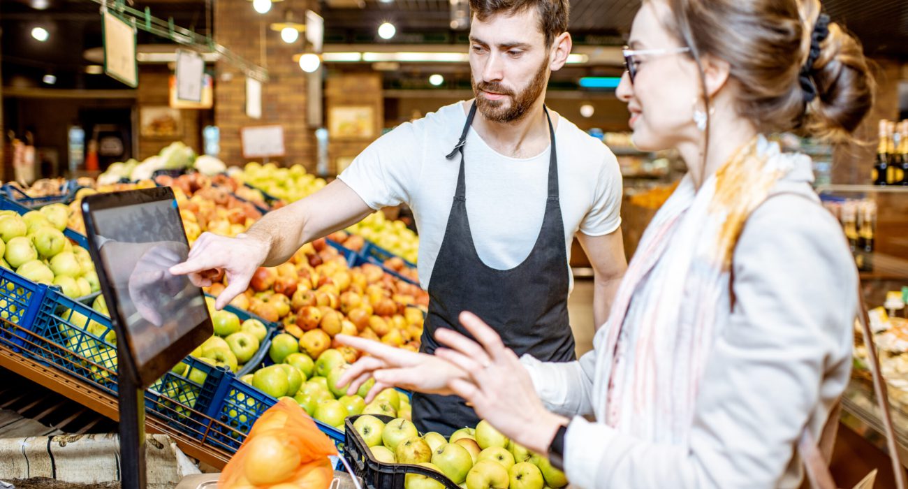 Handsome worker in uniform helping young woman customer to weigh apples on the scales in the supermarket